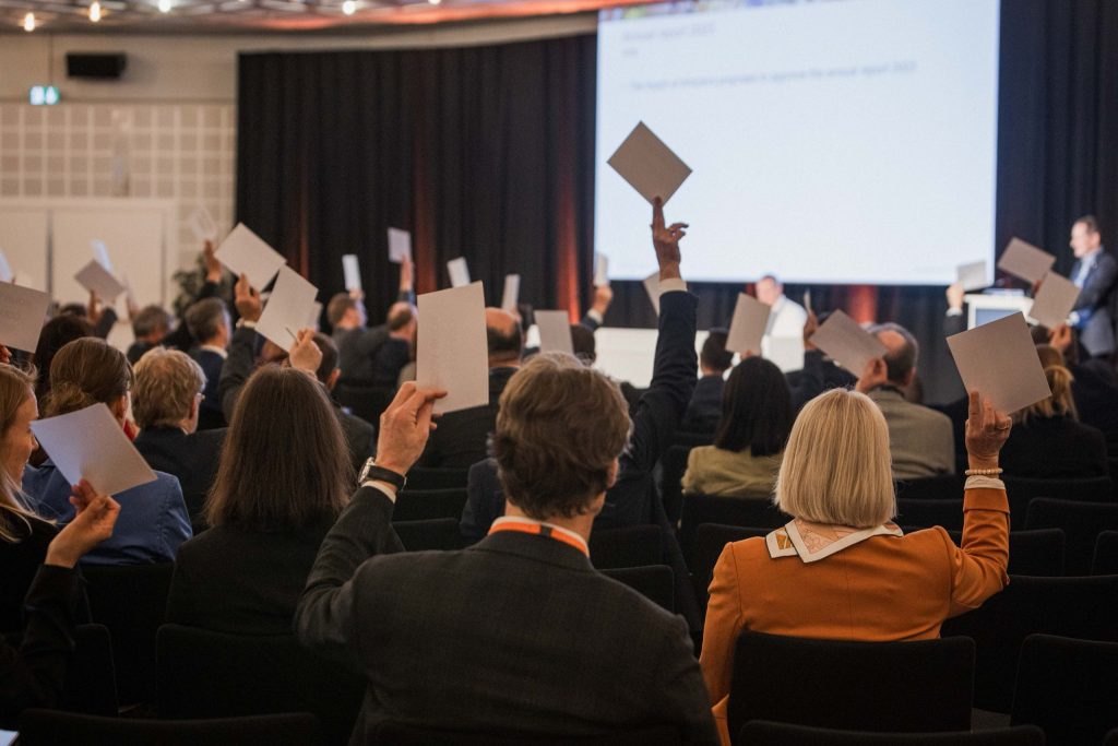 Members raising their voting cards at the Swiss Biotech Association general assembly 2024, the agenda on the screen ahead
