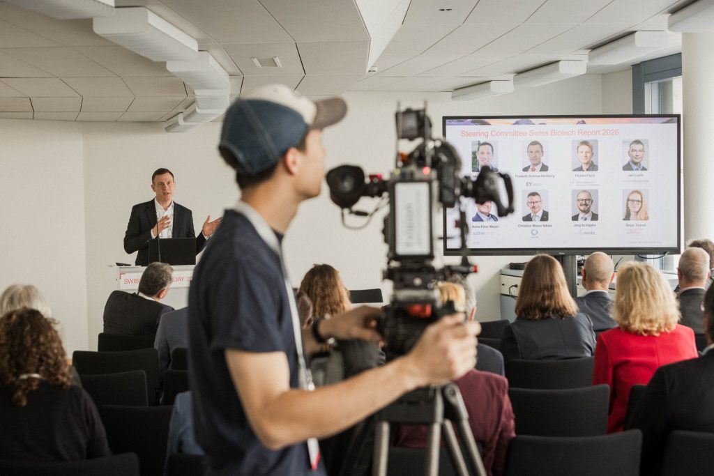 A speaker presenting the steering committee of the Swiss Biotech Report 2026 at the media conference of the Swiss Biotech Day 2026, a camera operator in the foreground