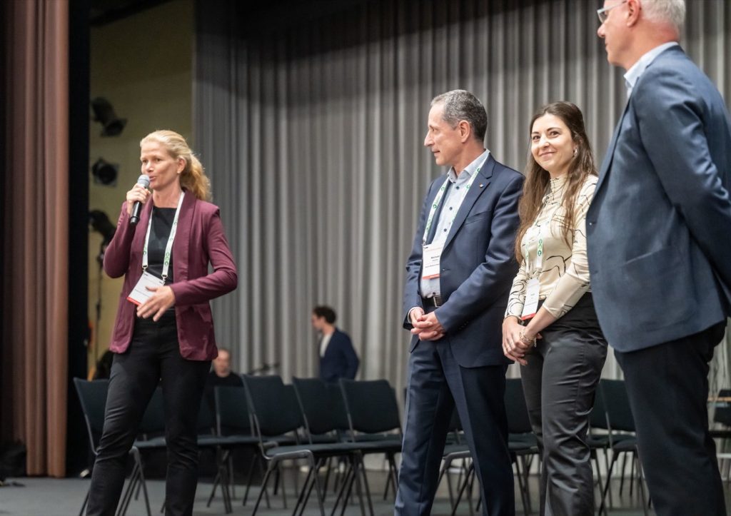 Speaker with a microphone beside the stage, three panelists standing next to her